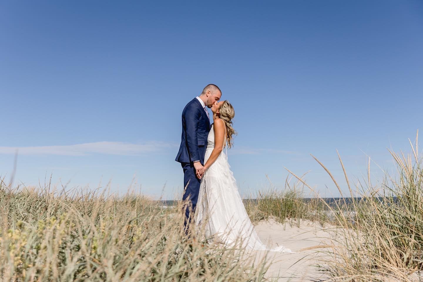 Bride and groom kissing among beach dunes under a clear blue sky